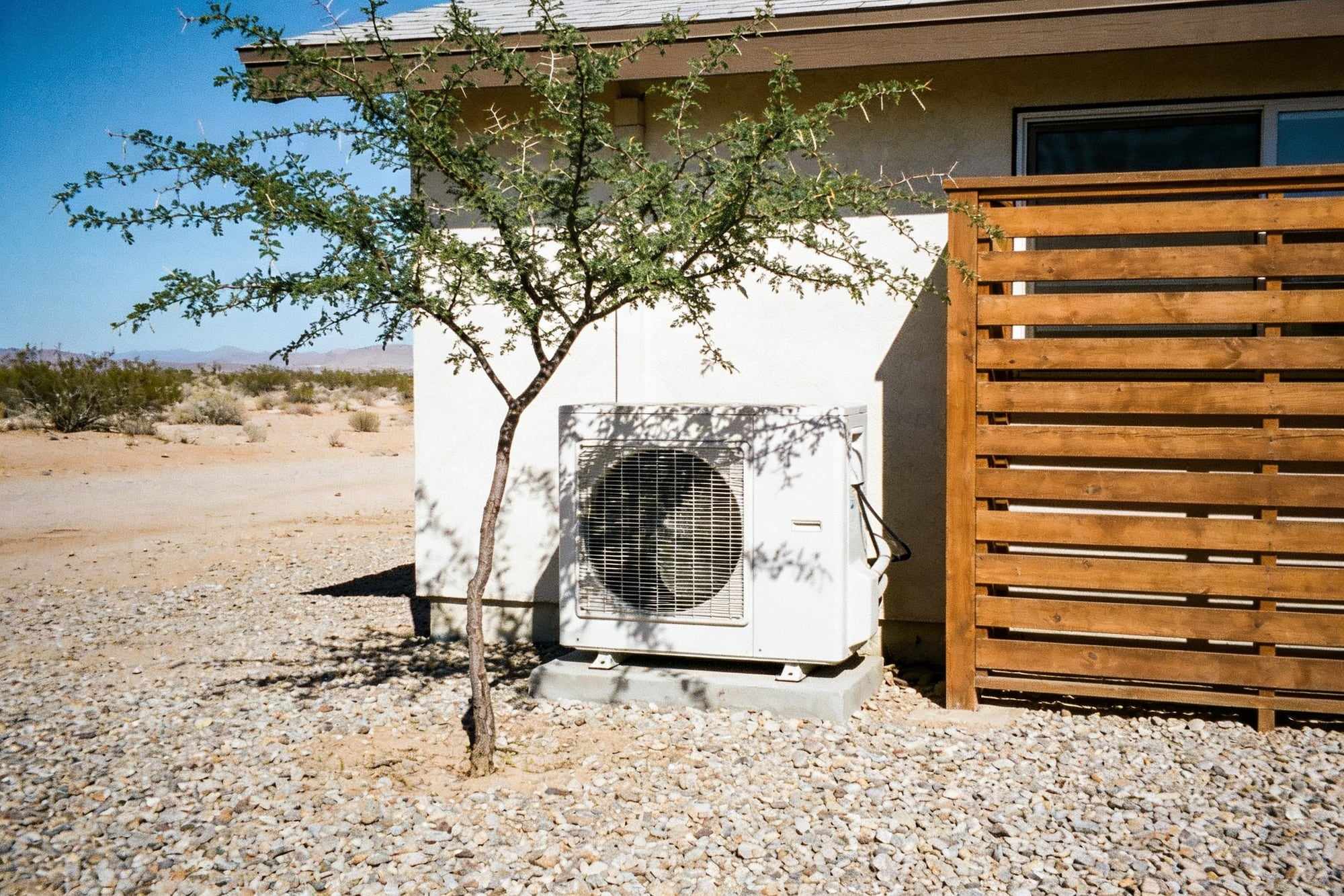 An outdoor AC unit shaded by a small tree and a slatted screen